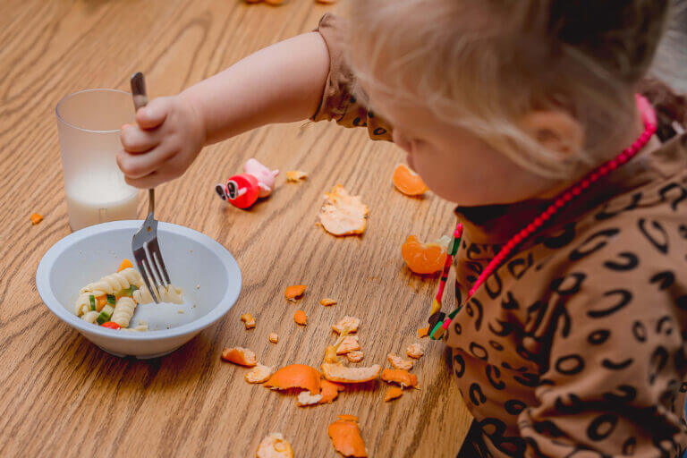 alt= A toddler practicing healthy eating by independently using a fork to eat a bowl of food in a child care kitchen.