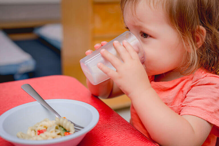 alt= A toddler practicing healthy eating by independently using a fork to eat a bowl of food in a child care kitchen.